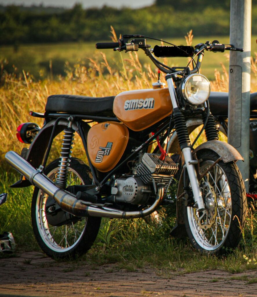 Image actuelle : Un cyclomoteur Simson orange, garé dans un champ, en lumière de fin de journée.