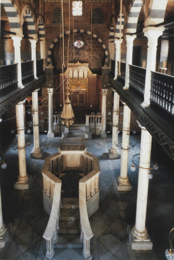 Intérieur d'une synagogue vu d'en haut. Au centre, une estrade octogonale en marbre clair, entourée de colonnes blanches et de galeries en bois sombre. Au fond, sous un arc à claveaux alternés noirs et blancs, une arche sainte dorée. Des lampes suspendues descendent du plafond.
